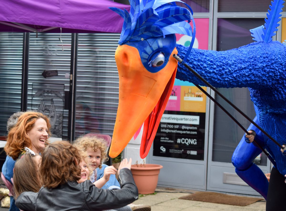 Giant blue bird puppet appears to bite audience members at an outdoor theatre performance