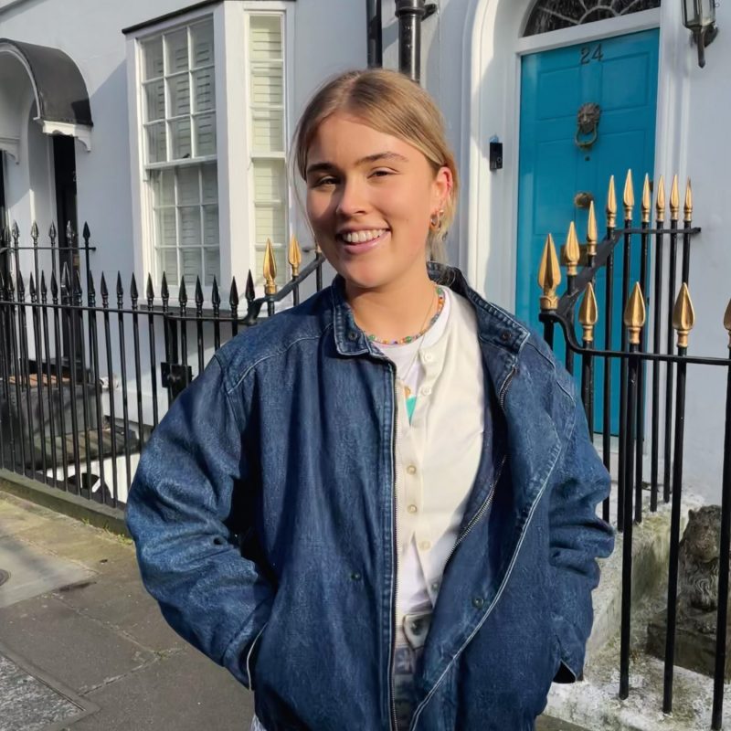 An image of Katie Inglis wearing a denim jacket, standing in front of a line of houses