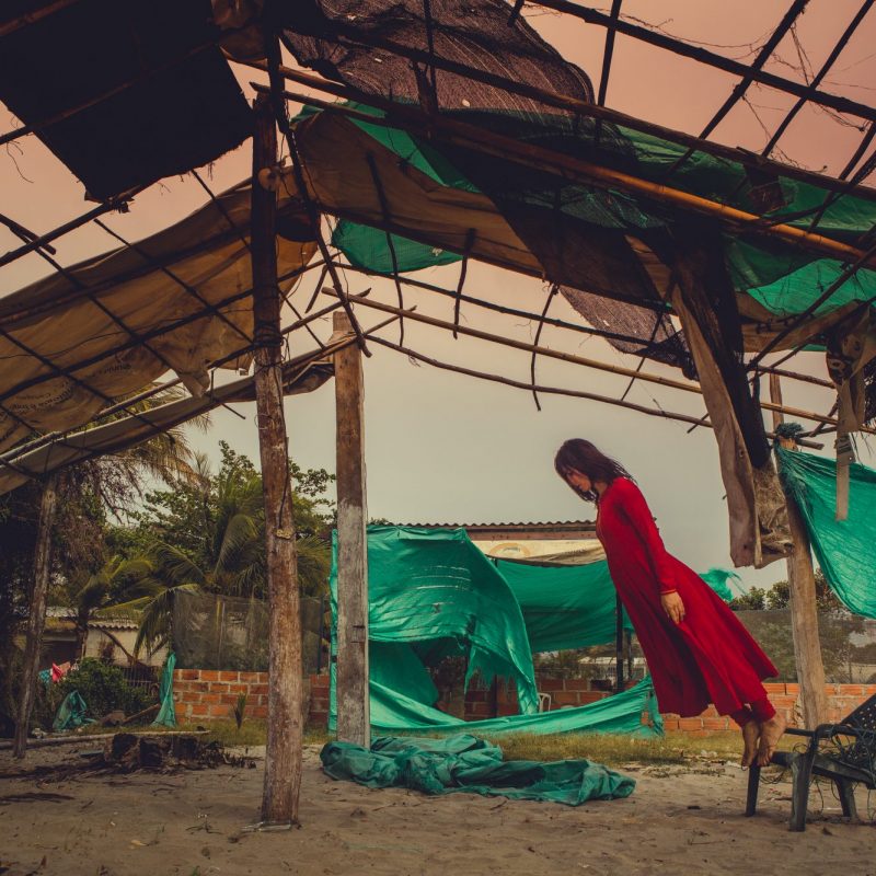 Woman in a long red dress standing on a small plastic chair, leaning forwards almost diagonally. She is standing under the framework of a house - like structure, with beams holding it up and green tarpaulin around the sides.