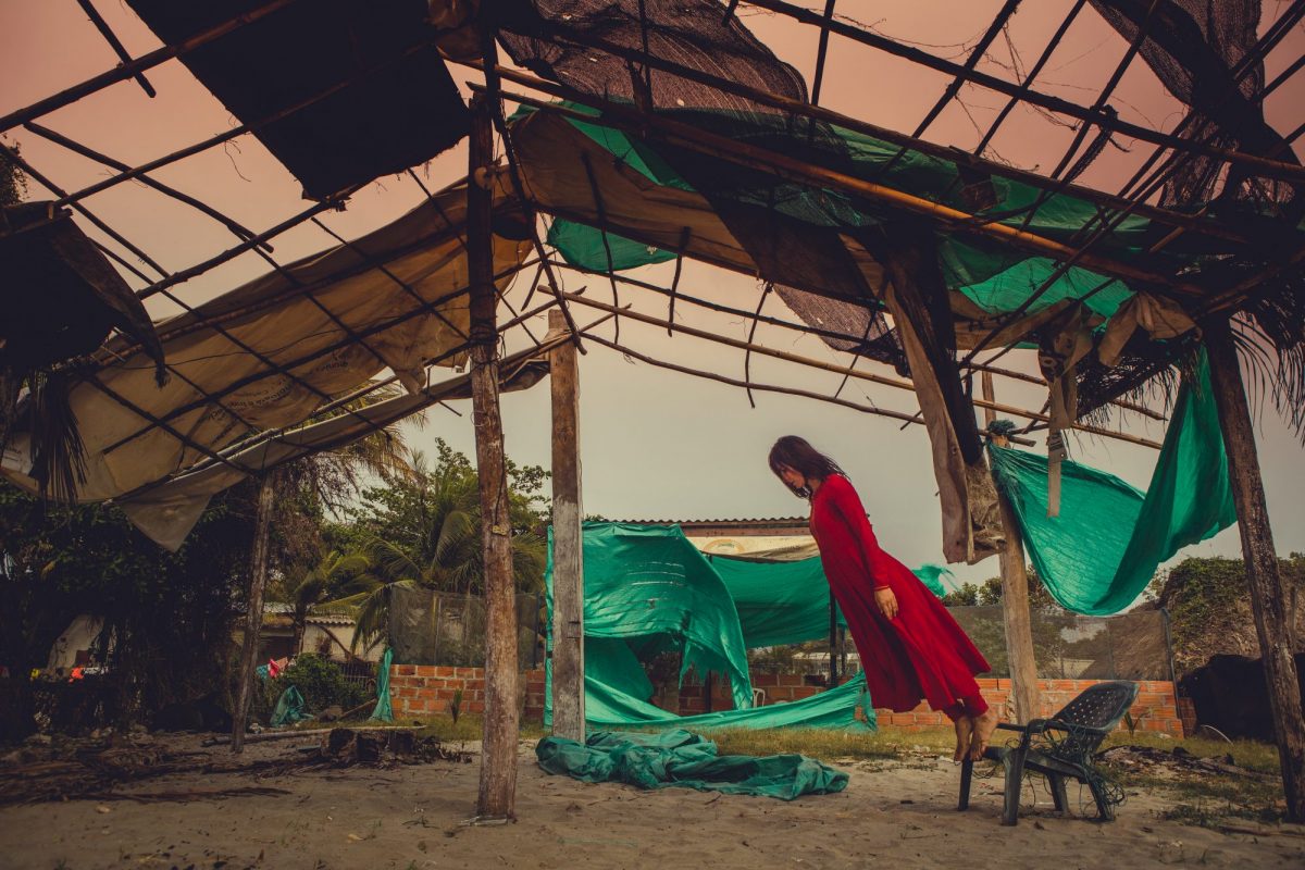 Woman in a long red dress standing on a small plastic chair, leaning forwards almost diagonally. She is standing under the framework of a house - like structure, with beams holding it up and green tarpaulin around the sides. 