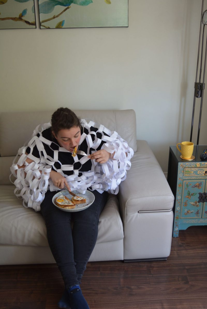 A real-life photograph depicts a girl with dark hair, sitting on a cream leather sofa, eating fried egg on toast. She is covered in white paperchains all over her body. The floor is a dark wooden colour and the wall behind the sofa is off-white.