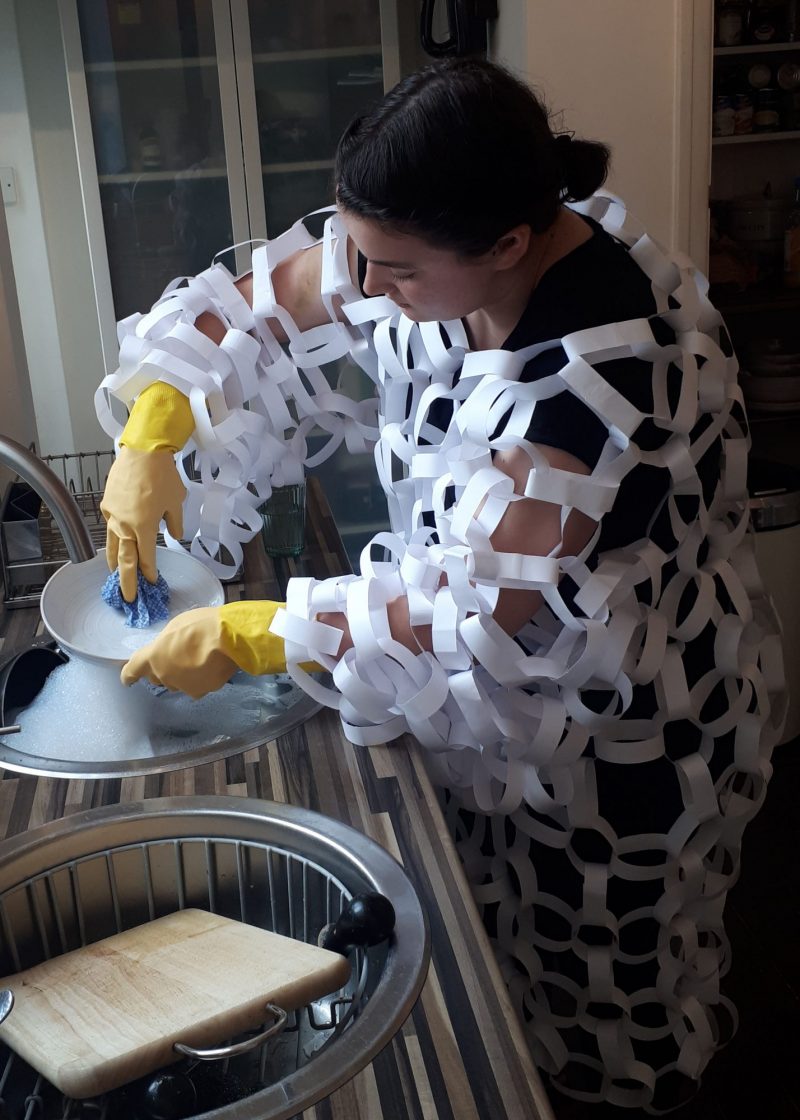 A real-life photograph depicts a girl with dark hair at a sink washing up a plate, with yellow washing up gloves on. She is covered in white paperchains all over her body. She looks down at the plate she is washing.