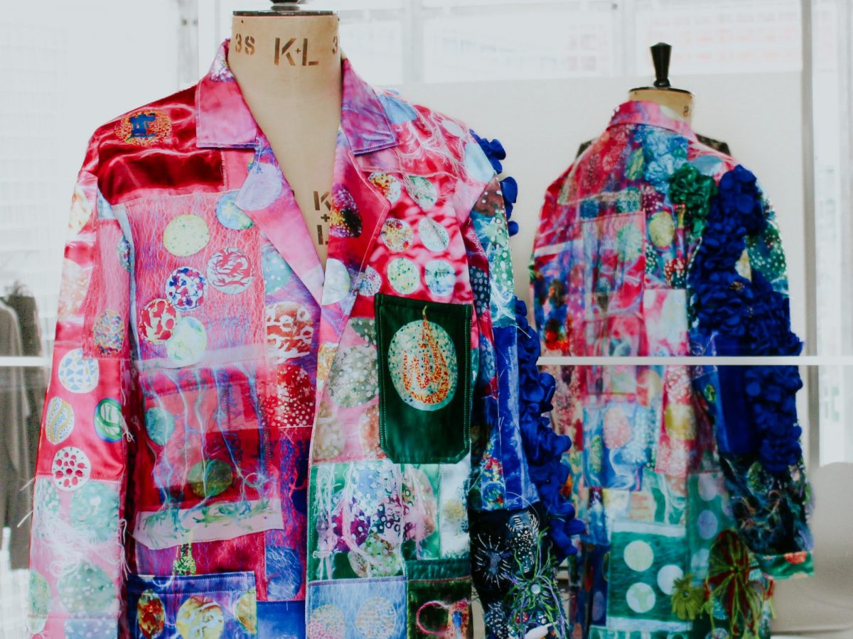 A multi-coloured lab coat made up from multiples textile patches, each decorarted with a design. It is reflected in a mirror.