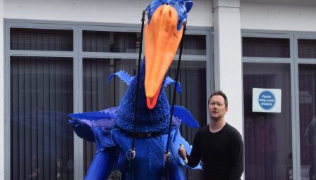 Izzy wearing a giant bird-like puppet, with Phil Pearson