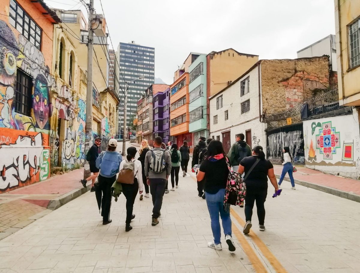 A group walk up a street in Bogota, Columbia. The buildings are multi-coloured and there is a lot of street art.