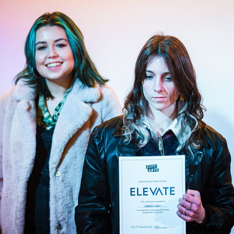 Young artist holds certificate reading ELEVATE. They are standing next to the course leader.