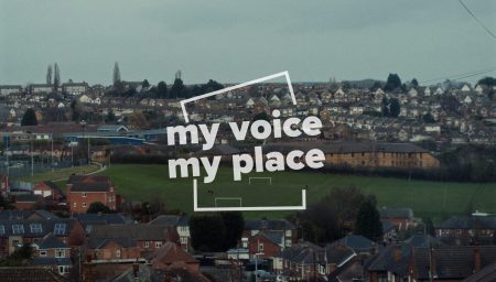 A panoramic view of a suburban landscape in Carlton, England, overlaid with the white, translucent text 'my voice my place'. The image shows rows of houses with varying roof designs on a hillside, a green football field in the mid-ground, and leafless trees scattered throughout, against a subdued, overcast sky.