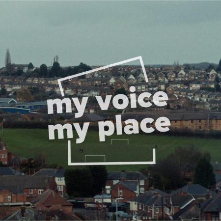 A panoramic view of a suburban landscape in Carlton, England, overlaid with the white, translucent text 'my voice my place'. The image shows rows of houses with varying roof designs on a hillside, a green football field in the mid-ground, and leafless trees scattered throughout, against a subdued, overcast sky.