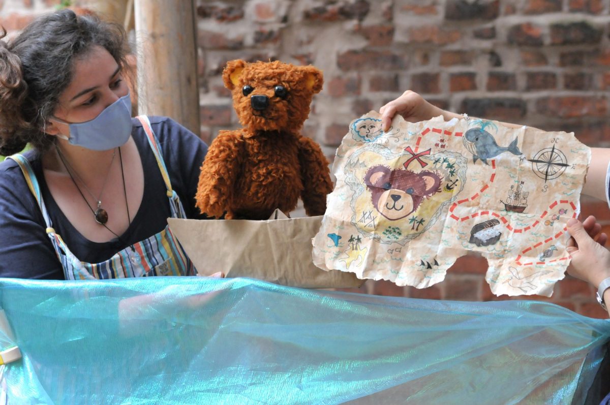 A person engaged in creative play displays a colorful, roughly drawn map to a teddy bear, complete with maritime and animal illustrations, all set in a workshop with a brick wall backdrop.