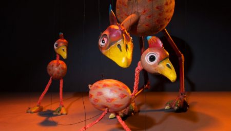Whimsical bird marionettes with vibrant colors and intricate textures suspended by strings against an orange backdrop.
