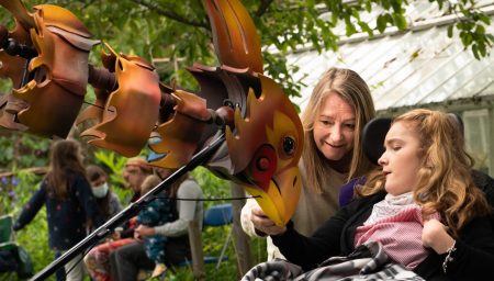 A tender moment as a smiling woman leans in to share a colorful bird puppet with a young girl in a wheelchair, both engrossed in the puppet's details, in a lush garden setting.