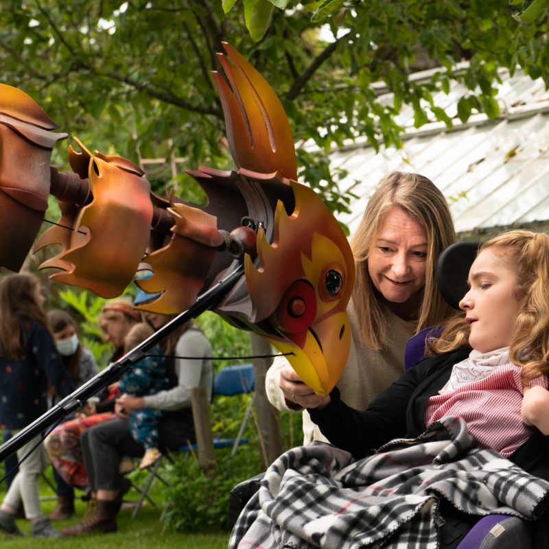 A tender moment as a smiling woman leans in to share a colorful bird puppet with a young girl in a wheelchair, both engrossed in the puppet's details, in a lush garden setting.