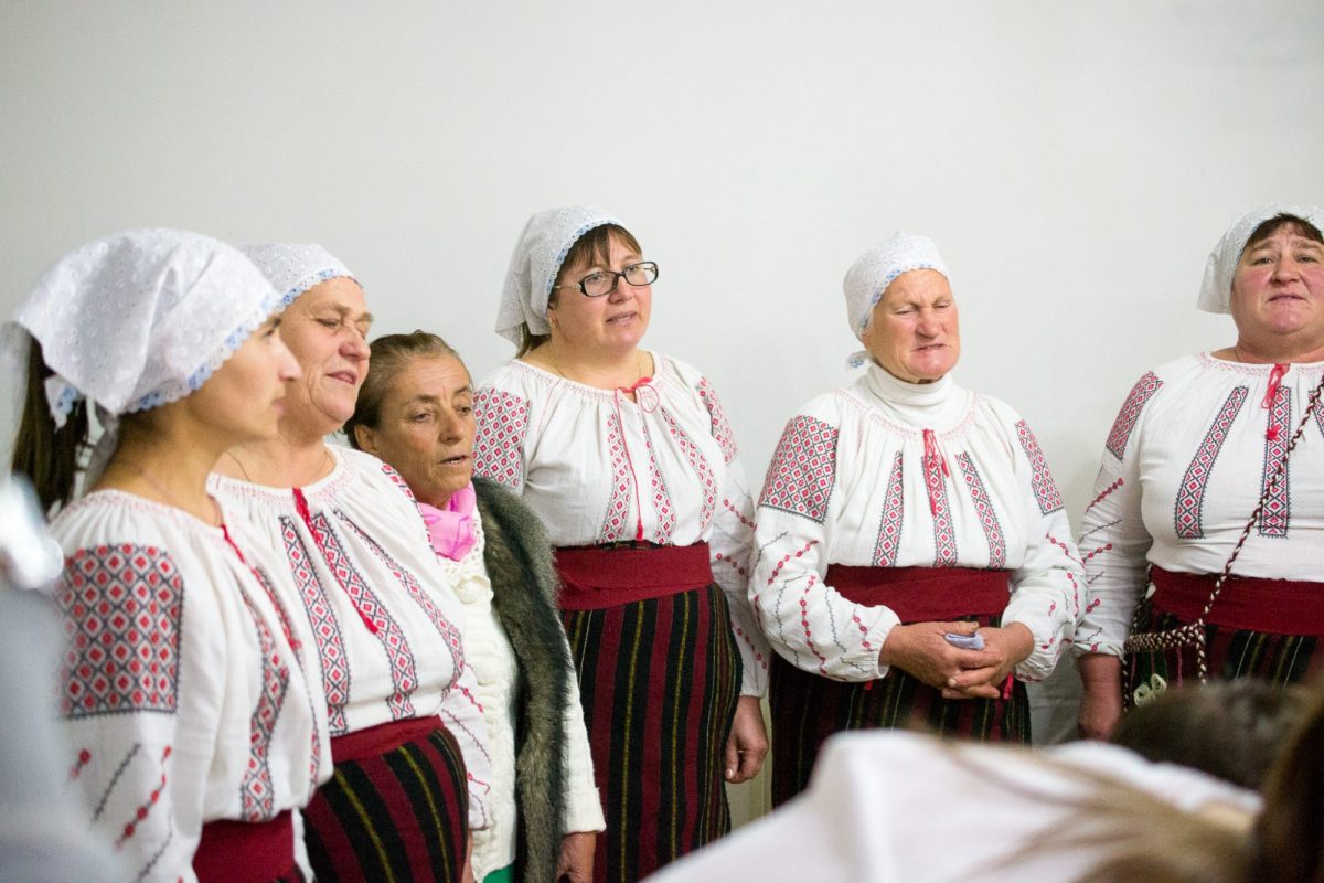 A group of women in traditional embroidered dresses and headscarves singing at a cultural event. 