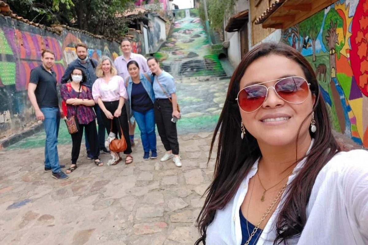 A selfie of a smiling woman with sunglasses in the foreground, with a group of people posing in the background on a colourful, mural-decorated street.