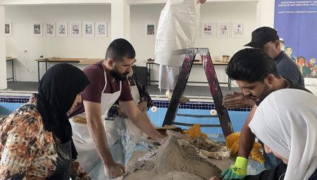 In a well-lit art studio, a woman in a white hijab and apron stands on a ladder overseeing a group of focused artists shaping a substantial clay piece on a table.