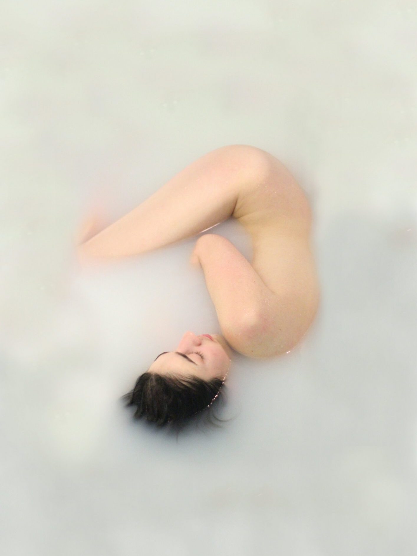 A person is submerged in milky water, resting in a fetal position with only their face and knees emerging from the surface.