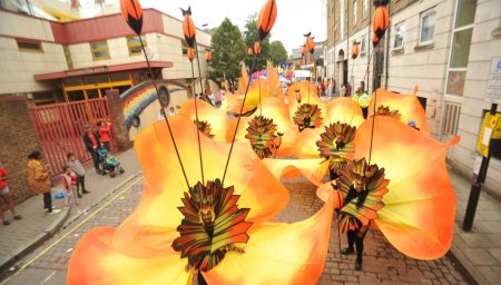 Performers in vibrant orange flower costumes parade down a street at a festive event.