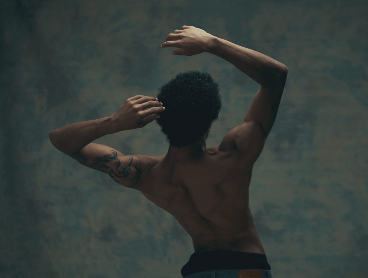 A shirtless man with a tattoo on his upper arm stands with his back to the camera, his arms raised and crossed over his head in a graceful, dance-like pose.
