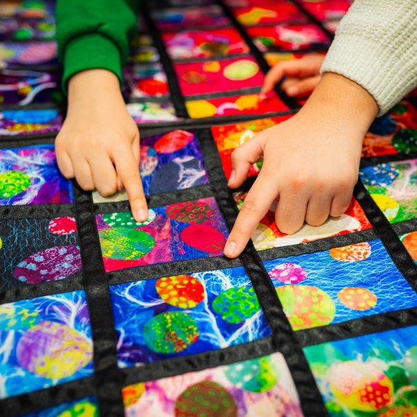 Close-up of two children's hands pointing at colorful, patterned squares on a quilted mat, showcasing a variety of textures and vibrant hues.