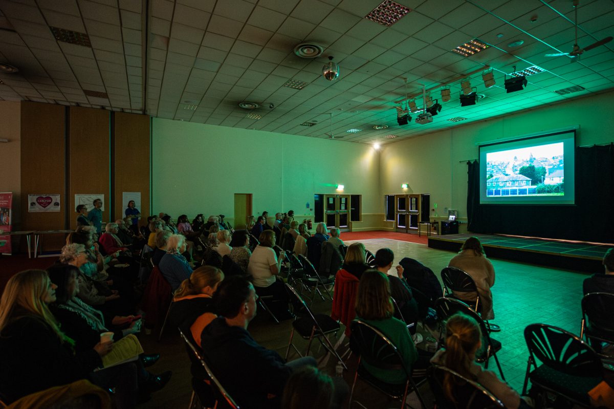 A diverse audience in a dark auditorium watches a presentation, focused on a brightly lit projection screen displaying an image of a traditional housing estate.