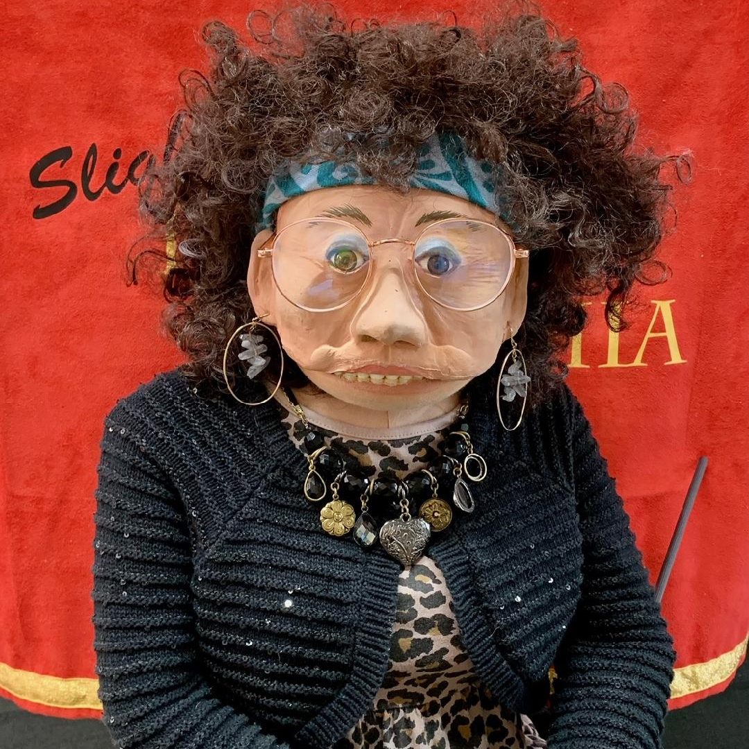 A characterful puppet with a detailed, expressive face is styled with curly hair, oversized round glasses, and a fashionable ensemble. The puppet wears a leopard print blouse, a knit cardigan, and is adorned with large earrings and multiple necklaces. She is seated before a luxurious red velvet background with the text 'Slightly Psychic Cynthia' in elegant script.