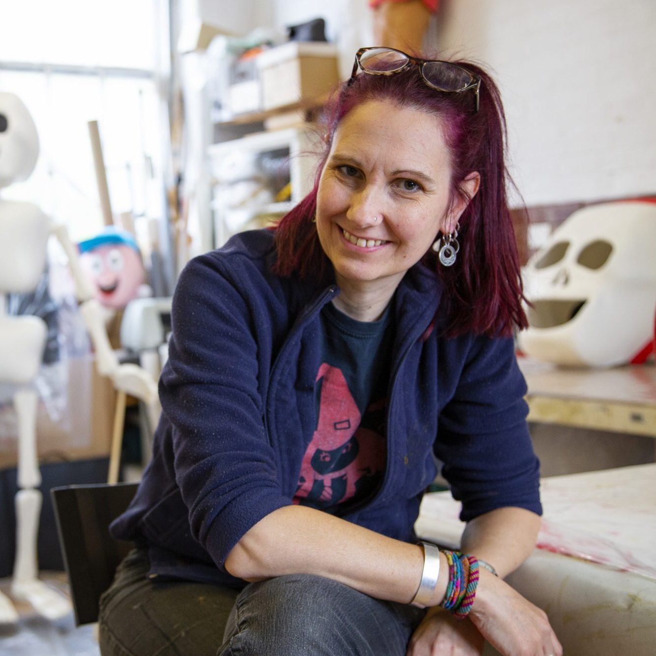 A woman with vibrant red hair and a warm smile sits casually in a puppet-making workshop. Behind her, an array of whimsical puppet figures with exaggerated skull-like faces are perched, showcasing different stages of the creative process. She wears a dark jacket over a playful t-shirt, and her sunglasses are perched atop her head, suggesting a lively and artistic work environment.