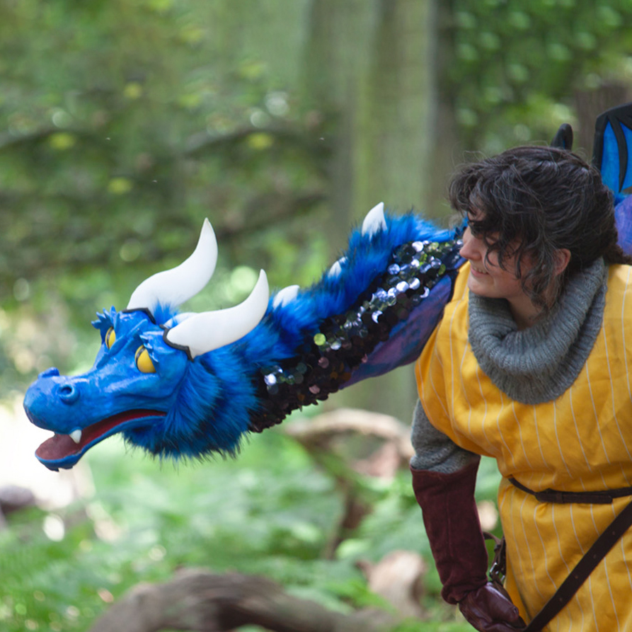 A person in a medieval costume interacts with a vibrant blue dragon puppet. The dragon, adorned with sequins and sporting prominent white horns, seems to be playfully engaging with the individual, who is smiling and looking towards the creature with a sense of wonder. The setting is outdoors with a backdrop of lush green foliage.