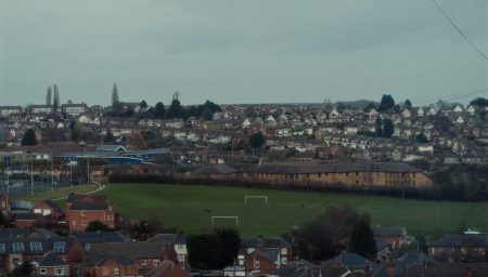 A sweeping view of Carlton, England, with densely packed residential houses and buildings. The foreground shows rows of homes with reddish-brown rooftops, while the background reveals a hill crested with rows of similar houses. A green football pitch is visible amidst the urban landscape, and the overall scene has a muted color palette, suggesting an overcast day.