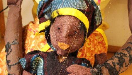 A detailed close-up of a handmade puppet depicting an African woman. The puppet is dressed in a vibrant, patterned fabric with blue, red, and yellow colors and wears a matching headwrap adorned with a yellow band. The face features stitched eyes, a nose, and a mouth, creating a serene expression.