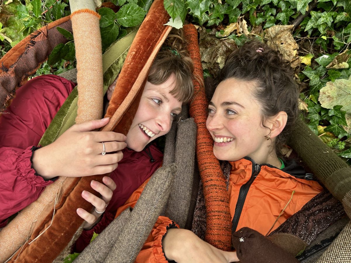 Two smiling women lying down surrounded by various textured and colored fabric tubes, resembling large plant stems or vines. Both are wearing jackets, one in red and the other in orange. They are looking at each other affectionately, with a background of green foliage and fallen leaves.