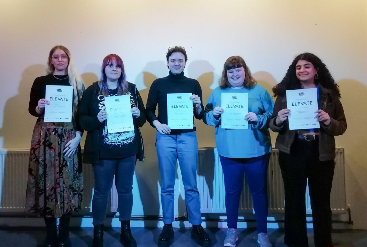 A group of five individuals standing in a row, each holding a certificate labeled 'ELEVATE'. They are standing against a plain background and smiling for the camera.