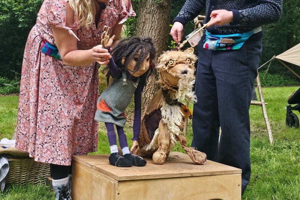 Two puppeteers performing outdoors in a grassy area with trees in the background. The female puppeteer on the left, wearing a floral dress, is operating a puppet of a child with dark hair and a gray outfit. The male puppeteer on the right, wearing a dark sweater, is manipulating a puppet resembling a dog. Both puppets are positioned on a wooden box.