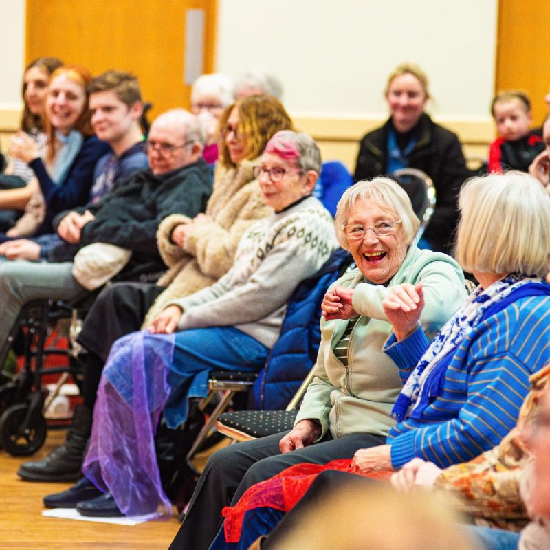 An audience seated in rows, watching a performance or presentation. Many of the attendees are elderly, and they are engaged and smiling. The setting is a community hall with beige walls and bright lighting.