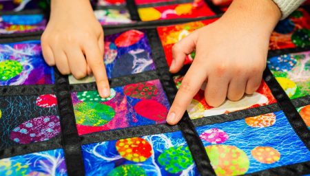 Close-up of two children's hands pointing at a colorful patchwork. The textile features a variety of bright and patterned squares, each with unique designs and textures, creating a vibrant mosaic effect.
