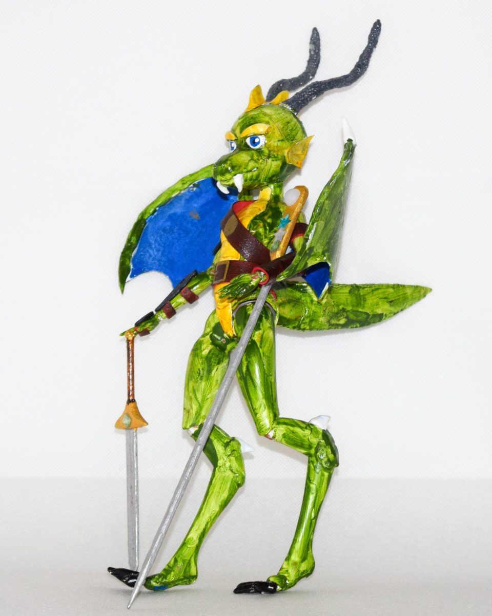 A colourful, handmade model of a fantasy dragon-like creature standing upright. The figure is painted bright green with yellow accents, large blue eyes, and long black horns. It has blue wings, clawed feet, and carries two long spears, one in each hand. A brown strap crosses its chest, and its expression appears fierce yet playful. The background is plain white, making the model stand out clearly.