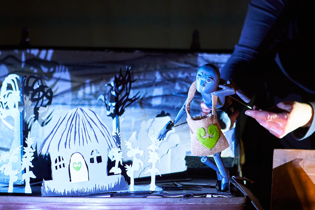 A grey wooden puppet with large eyes and a hessian bag marked with a green heart is being manipulated on a small stage. In front of it are paper cut-out trees and a house with a green heart on the door, lit in bright white and blue light.