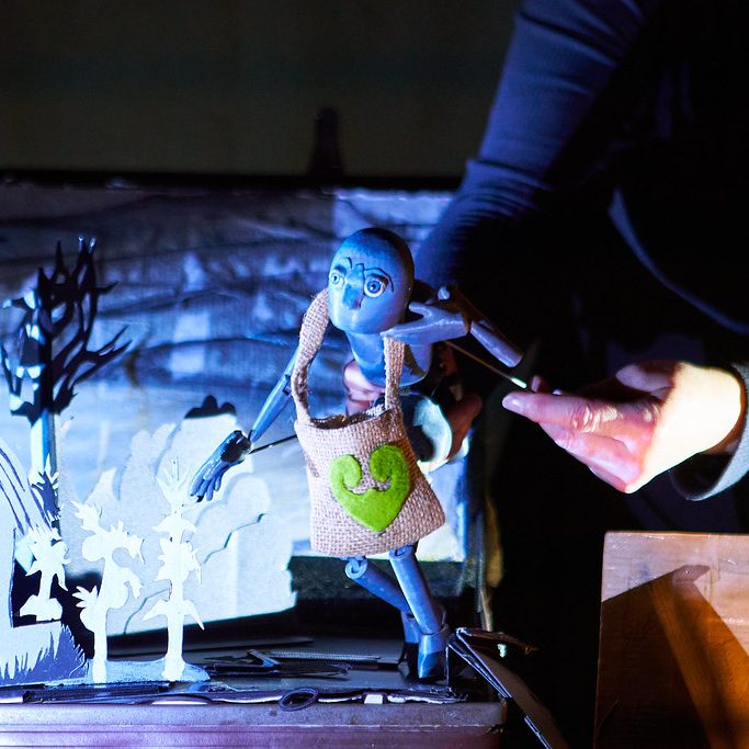 A grey puppet with a green heart on its chest peeks from a miniature stage, manipulated by a performer’s hands