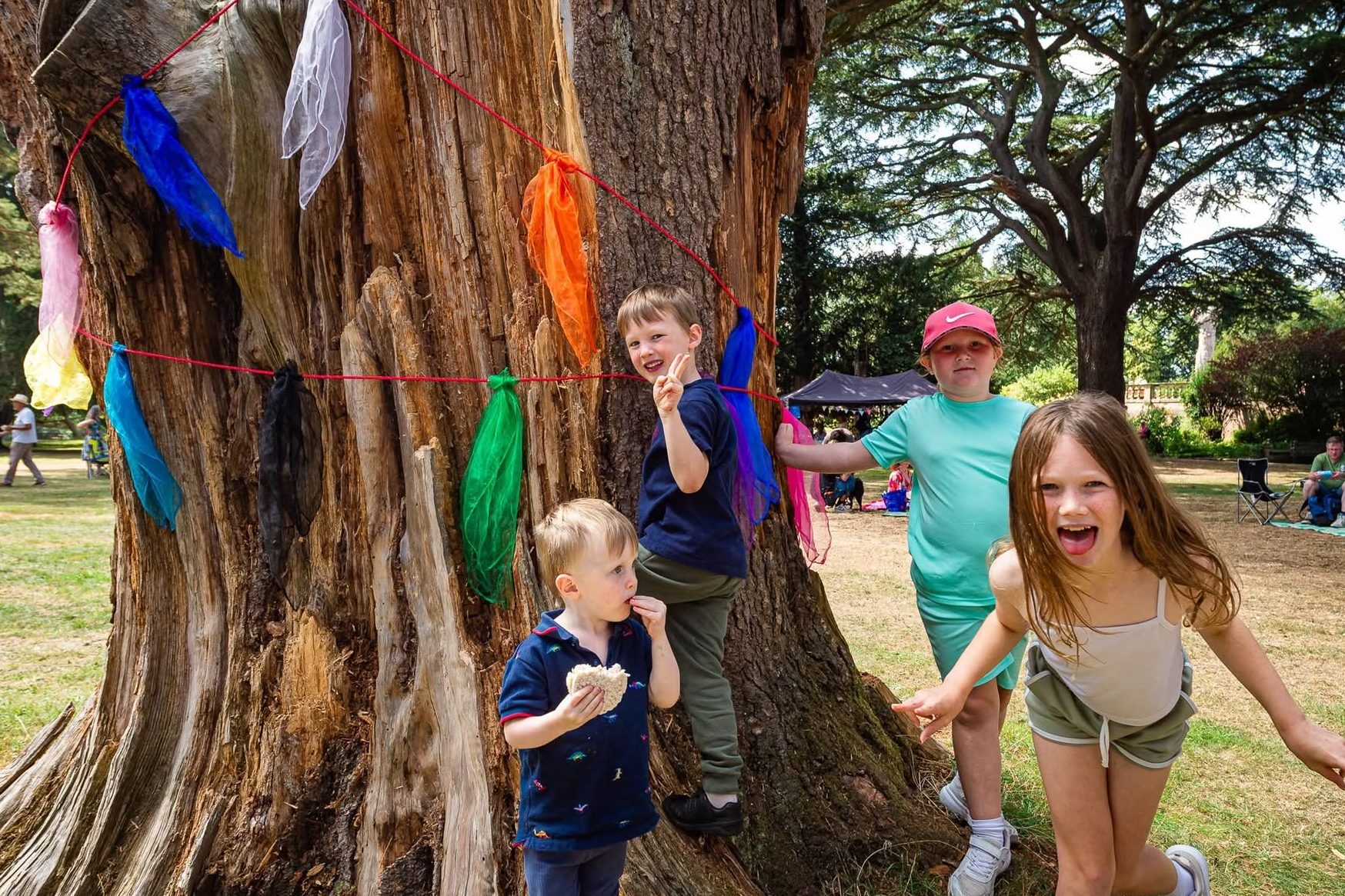 Four children play around the base of a large tree at an outdoor event. The tree trunk is decorated with a red string holding colourful scarves. The children are laughing and smiling, with one eating a sandwich. In the background, people are seated on picnic blankets and chairs under tall trees.