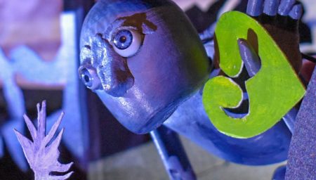 Close-up of a grey puppet figure with wide eyes, part of a theatrical set. The puppet has a large green leaf and is framed by cut-out foliage shapes, lit dramatically in purple and blue tones.