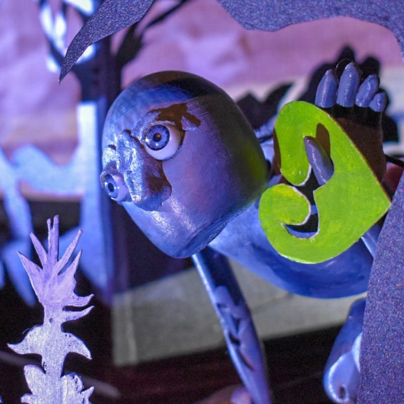 Close-up of a grey puppet figure with wide eyes, part of a theatrical set. The puppet has a large green leaf and is framed by cut-out foliage shapes, lit dramatically in purple and blue tones.