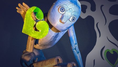 Promotional graphic for SeedHeart. A blue wooden puppet with large eyes holds a green heart shape against its chest, leaning forward as if in motion. Behind it is a stylised black and white tree with a green heart at its base. Bold white text reads: ‘SEEDHEART.’ Below, smaller text says: ‘Touring North Nottinghamshire Villages. A puppet show & workshop for ages 6+.’