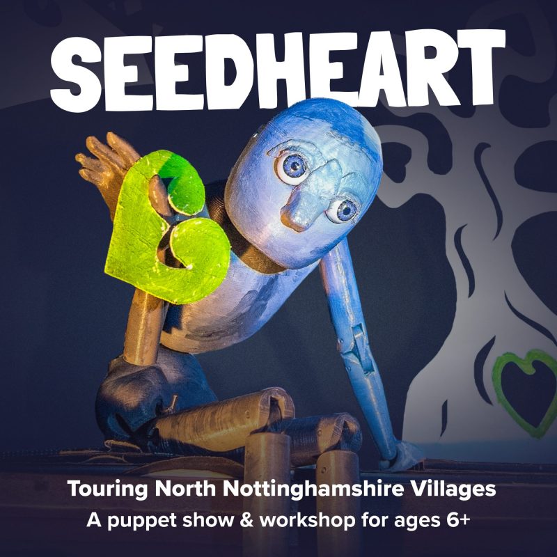 Promotional graphic for SeedHeart. A blue wooden puppet with large eyes holds a green heart shape against its chest, leaning forward as if in motion. Behind it is a stylised black and white tree with a green heart at its base. Bold white text reads: ‘SEEDHEART.’ Below, smaller text says: ‘Touring North Nottinghamshire Villages. A puppet show & workshop for ages 6+.’