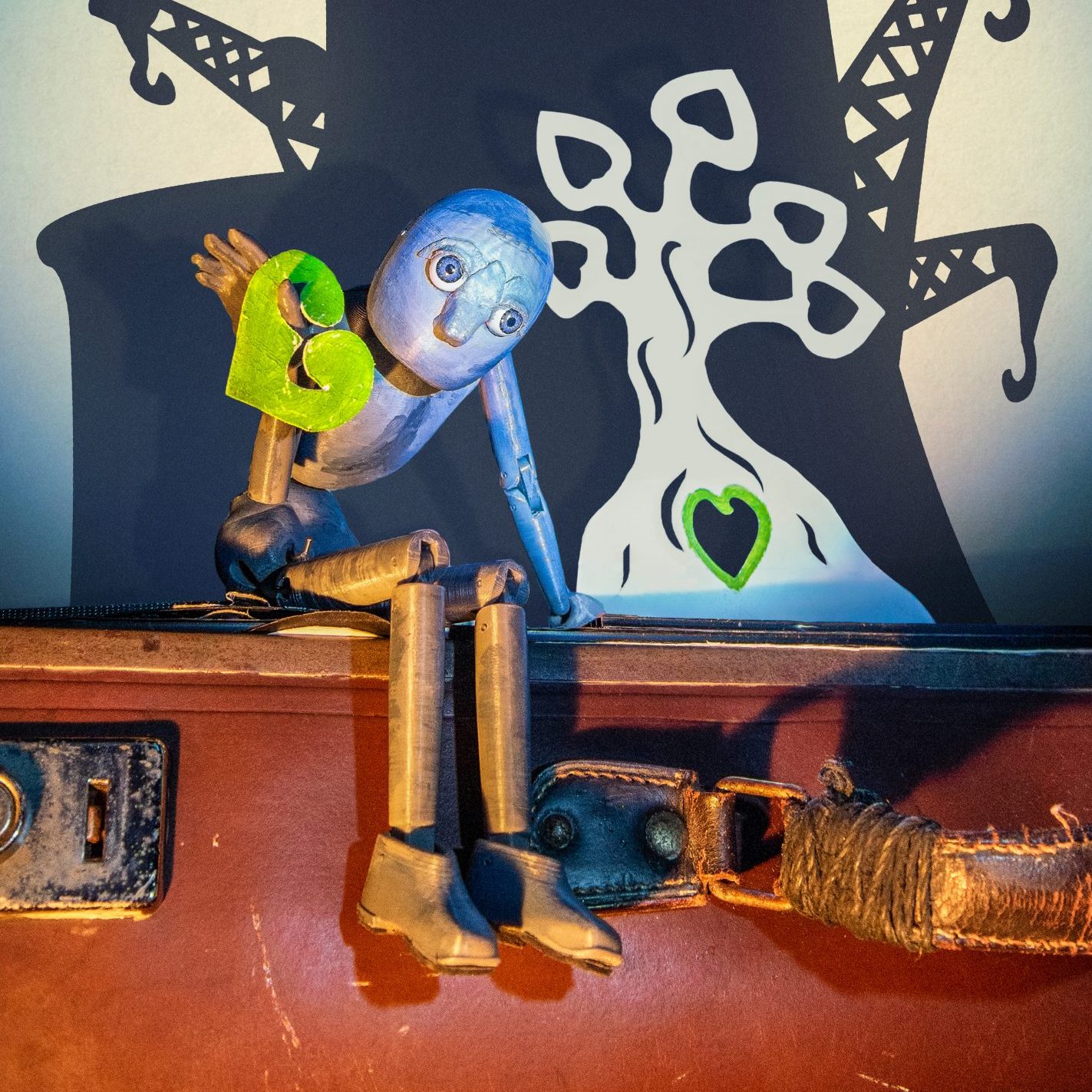 A grey puppet sits on the edge of a suitcase, holding a green heart. Behind it, a black-and-white cut-out tree with a heart-shaped hollow glows green