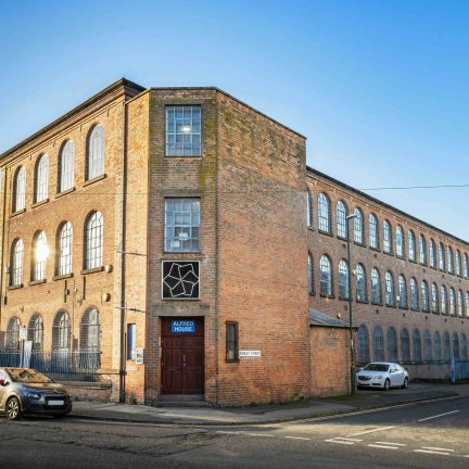 Exterior photograph of Alfred House, a large red-brick industrial building with rows of tall arched windows, located on Ashley Street in Nottingham. A blue sign above the door reads ‘ALFRED HOUSE’, with the BACKLIT logo mounted above. Cars are parked along the street beside the building under a clear blue sky.