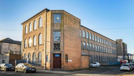 Exterior photograph of Alfred House, a large red-brick industrial building with rows of tall arched windows, located on Ashley Street in Nottingham. A blue sign above the door reads ‘ALFRED HOUSE’, with the BACKLIT logo mounted above. Cars are parked along the street beside the building under a clear blue sky.