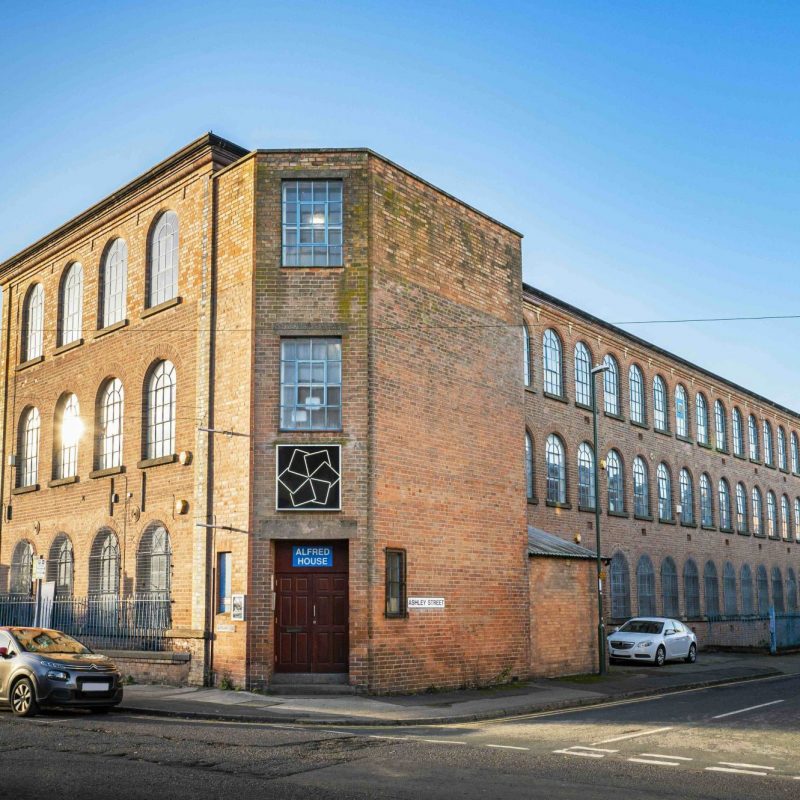Exterior photograph of Alfred House, a large red-brick industrial building with rows of tall arched windows, located on Ashley Street in Nottingham. A blue sign above the door reads ‘ALFRED HOUSE’, with the BACKLIT logo mounted above. Cars are parked along the street beside the building under a clear blue sky.