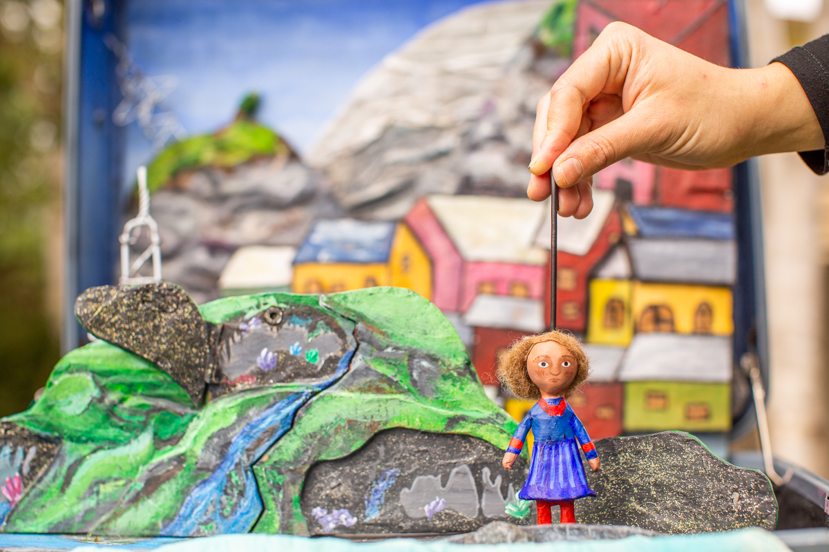 Close-up of a small handmade puppet of a girl in a blue dress being held upright by a thin rod. A hand controls the puppet from above. Behind it is a colourful miniature set with painted houses, green hills and a winding river.