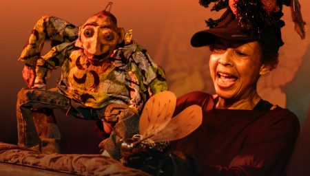 A performer animatedly operates a handmade puppet with large eyes and patterned clothing, leaning over a suitcase-style stage. Warm orange lighting fills the scene. The performer wears a decorative headpiece featuring an insect-like form, and holds a small winged prop in front of the puppet.