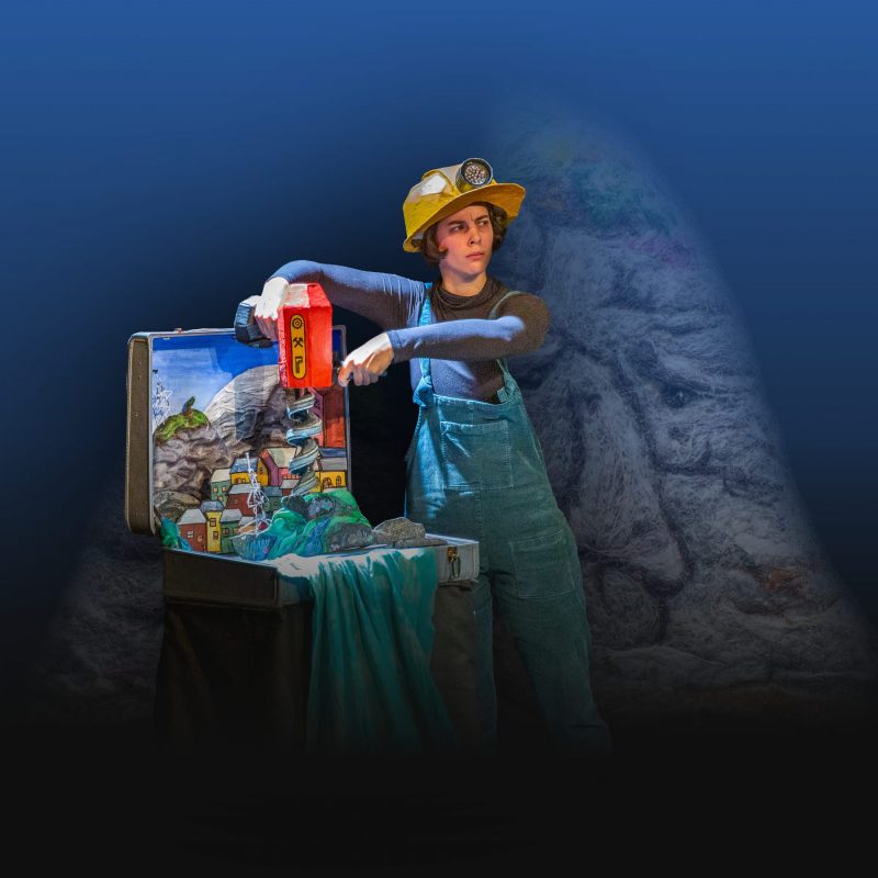 A performer wearing blue overalls and a yellow helmet with a headlamp stands beside an open suitcase set, operating a red drill-like prop over a miniature landscape of houses and rocks.