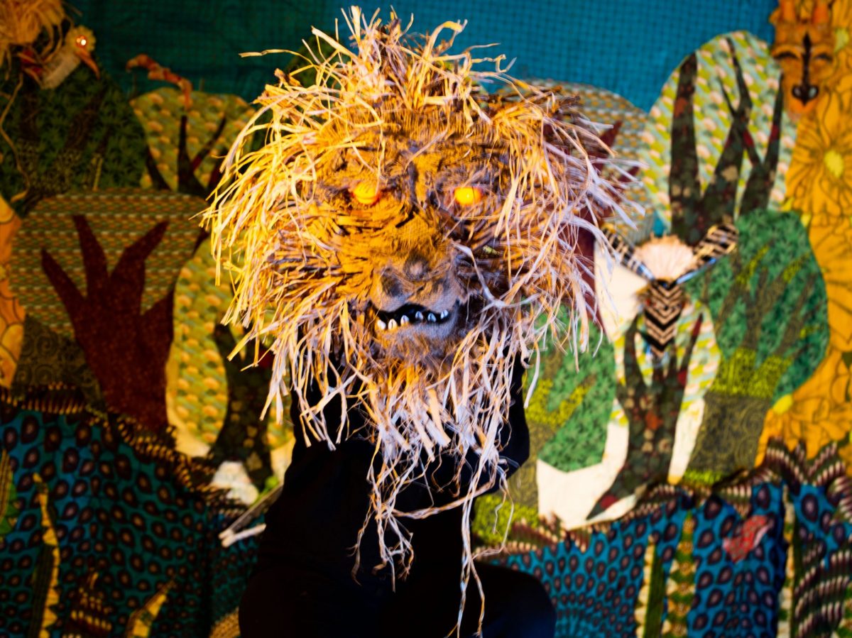 A large, expressive puppet head resembling a lion or wild creature with glowing orange eyes and a shaggy mane made from straw-like fibres is held up by a performer dressed in black.